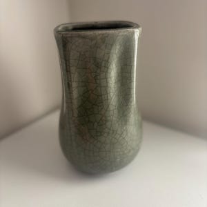 May include: A ceramic vase with a glossy, sage green glaze featuring a distinct crackle pattern across its entire surface. The vase has a slightly curved, hourglass-like silhouette with a wide, rounded base and a rectangular opening at the top.