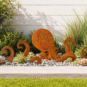 May include: A rust-colored metal octopus sculpture featuring a large, rounded head with textured circular patterns and multiple curved tentacles extending outward, positioned among small gray stones and green garden plants.