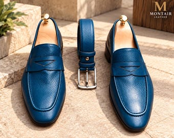 May include: A pair of blue textured leather penny loafers with wooden shoe trees and a matching blue leather belt with a silver buckle. The top right corner features the text M MONTAIR LEATHER in a gold-colored font on a dark background.