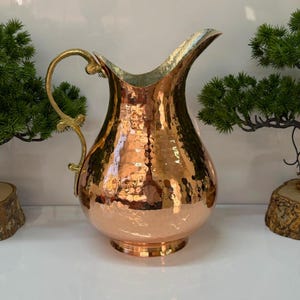 May include: A hammered copper pitcher with a wide, rounded body and a narrow neck. The pitcher features a polished, reflective surface and a decorative, curved brass handle attached to the side. The rim is flared, creating a distinct pouring spout.