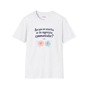 May include: A white short-sleeved t-shirt featuring the dark blue text Are you an assertive or an aggressive communicator? above two speech bubbles, one pink with a heart and one light blue with a lightning bolt, and a small purple heart.