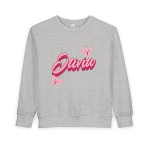May include: A heather gray long-sleeve sweatshirt featuring the name Olivia printed in a bold, dark pink script font. The design is accented with two light pink butterflies, one positioned above the text and one below the text on the left.