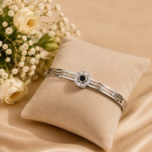 Può includere: Un bracciale rigido in metallo color argento con un elemento circolare centrale caratterizzato da una pietra nera circondata da piccoli cristalli trasparenti. Il bracciale è composto da tre fili paralleli con piccoli accenti a rombo.