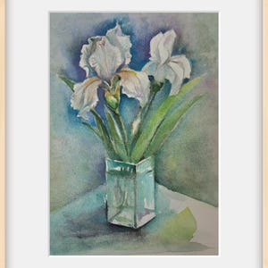 Può includere: Un dipinto ad acquerello raffigurante due fiori di iris bianchi con steli e foglie verdi in un vaso di vetro quadrato. Lo sfondo presenta lavaggi sfumati di toni blu, viola e verdi. L'opera è presentata all'interno di una cornice in legno chiaro.