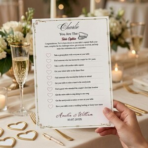 May include: A white rectangular wedding table game card titled Charlie You Are The Table Captain with a list of ten tasks to complete, featuring heart checkboxes, decorative gold borders, and the names Amelia & William with the date 10.07.202X.