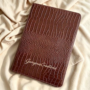 May include: A rectangular notebook with a dark brown cover featuring a textured crocodile skin pattern. The front cover displays the text Geergous Cractions in a gold cursive font. The notebook has rounded corners and a structured, firm exterior.