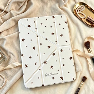 May include: A rectangular white tablet case featuring a pattern of small brown stars and segmented folding lines. The brand name Geo.Creations is printed in a dark script font near the bottom center of the cover, which has a smooth finish.