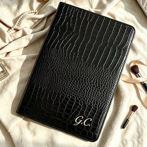 May include: A rectangular black notebook with a textured crocodile skin pattern. The cover features the gold-coloured initials G.C. embossed in the bottom right corner. The notebook has a clean, structured design with rounded corners.