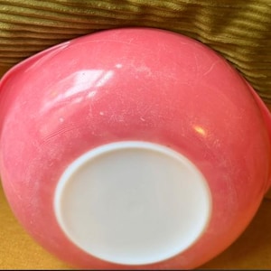 May include: A round mixing bowl with a vibrant pink exterior and a smooth, solid white circular base. The bowl features a curved rim with a small pouring spout on one side. The surface of the pink plastic shows light signs of wear and fine scratches.