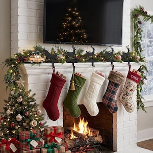 May include: Five Christmas stockings hang from black metal reindeer stocking holders on a white brick fireplace mantel. The stockings include a solid red knit, a solid green velvet, a white cable knit, a plaid, and a cream embroidered stocking.