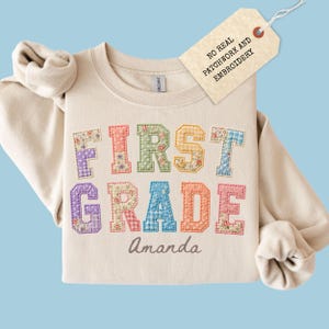 May include: A beige crewneck sweatshirt featuring the text FIRST GRADE in multi-colored, patterned block letters with a stitched appearance. Below the text, the name Amanda is printed. A tag attached reads NO REAL PATCHWORK AND EMBROIDERY.