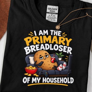 Puede incluir: Una camiseta negra con el gráfico de una patata marrón descansando en un sofá azul oscuro, sosteniendo un mando y una bebida roja. El texto dice I AM THE PRIMARY BREADLOSER OF MY HOUSEHOLD, con un cuenco de patatas fritas amarillas.