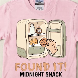 Peut inclure: Un t-shirt rose clair avec un motif de réfrigérateur ouvert contenant une bouteille de lait, un pot et un morceau de fromage. Une main attrape un cookie aux pépites de chocolat. Le texte indique FOUND IT! MIDNIGHT SNACK.