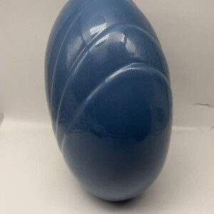 May include: A smooth, egg-shaped ceramic vase in a solid medium blue color. The surface features a series of raised, diagonal, curved ridges that wrap around the body of the vessel, creating a subtle texture and depth across the glossy finish.