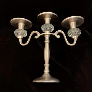 Può includere: Un candelabro a tre bracci realizzato in metallo color argento spazzolato, con uno stelo centrale e due bracci laterali curvi. Ogni braccio è decorato con una sfera in vetro trasparente e scanalato sotto un piattino circolare piatto.