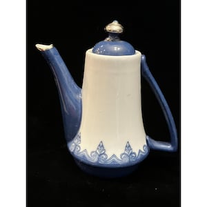 May include: A ceramic teapot featuring a white body with a blue base, handle, and spout. The base is decorated with a blue and white scroll pattern. The lid is blue with a silver-colored knob. The teapot has a tall, cylindrical shape.