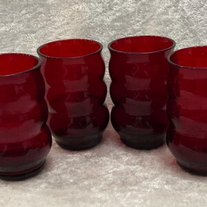May include: A set of four matching drinking glasses in a deep ruby red color. Each glass features a distinct rounded, bulbous silhouette with three horizontal ridges along the body, creating a textured, wavy profile that tapers toward the base.