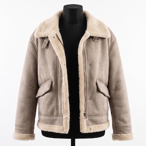 Könnte beinhalten: Eine beige Jacke mit cremefarbenem Kunstfellfutter, ausgestattet mit einem breiten Umlegekragen, langen Ärmeln mit Fellbündchen, zwei Vordertaschen mit Klappen, einem Frontreißverschluss und einem kleinen Schnallenriemen am Halsausschnitt.