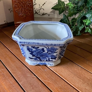 May include: A hexagonal ceramic planter featuring a white base with blue floral patterns and decorative border detailing. The vessel has a wide opening and a flared rim, with a small footed base, showcasing a classic blue and white color scheme.