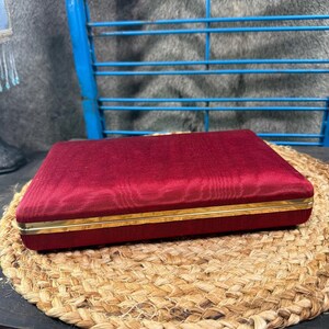 May include: A rectangular, deep red velvet-covered box featuring a thin, polished gold-tone metal trim along the center seam. The box has a smooth, fabric-textured exterior and a structured, flat-top design with clean, sharp edges and corners.