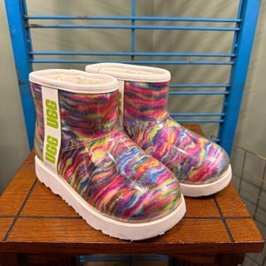 May include: A pair of ankle boots featuring a clear outer shell over a vibrant, multi-colored tie-dye pattern. The boots have a white vertical stripe on the side with the text UGG printed in lime green, and a thick white rubber sole.