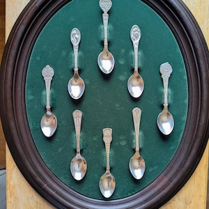 May include: A collection of seven silver-toned metal spoons with varied decorative handle designs, arranged in a circular pattern on a dark green velvet-lined oval display board with a dark brown wooden frame, showcasing intricate metalwork.