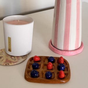 Pode incluir: Uma vela branca canelada com etiqueta dourada Rituals, um vaso de cerâmica alto com riscas verticais cor-de-rosa e brancas e rebordo de contas azuis, e um tabuleiro de cerâmica castanho em forma de waffle com pequenas esculturas de morangos e mirtilos.