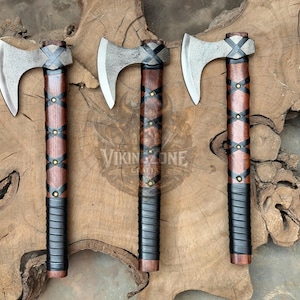 May include: Three axes with silver-toned metal heads and dark brown wooden handles. Each handle features a crisscross pattern of black leather straps secured with small brass-colored rivets and a black wrapped grip at the base. The text VIKING ZONE CRAFTS is visible.
