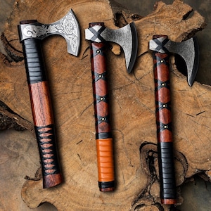 May include: Three axes with dark metal heads and wooden handles. The left axe features intricate silver-toned Norse-style engravings on the blade. The handles are wrapped in black and tan leather strips secured with small metal studs and bindings.