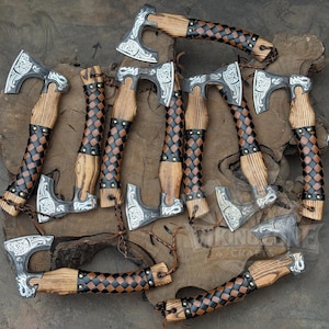 May include: A collection of hand axes featuring silver-toned metal heads with intricate engraved patterns and wooden handles wrapped in a black and brown checkered leather pattern, secured with small metal rivets along the grip.