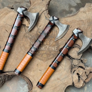 May include: Three hand axes with dark brown wooden handles wrapped in black leather straps and secured with metal rivets. The axe heads are made of textured grey steel with a curved blade edge. The handles feature a light brown grip section.