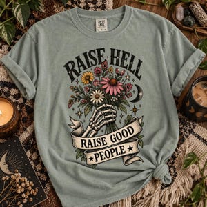 Könnte beinhalten: Ein salbeigrünes Kurzarm-T-Shirt mit einer Grafik einer Skeletthand, die einen Strauß bunter Blumen hält. Der Text RAISE HELL steht über den Blumen und RAISE GOOD PEOPLE auf einem Banner unter der Skeletthand, ergänzt durch Sterne und einen Mond.