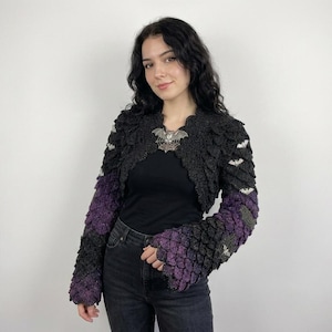 May include: A black and purple crocheted shrug featuring a textured scale-like pattern throughout the sleeves and body. The garment includes small white bat-shaped embellishments integrated into the design and a large silver bat brooch at the center.