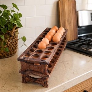 May include: A two-tiered rectangular wooden egg holder with a dark brown wood grain finish. The structure features multiple circular cutouts designed to securely hold individual eggs in place. Several light brown eggs are nestled in the top tier.