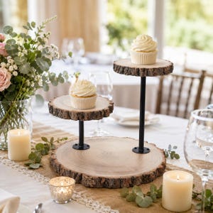 May include: A three-tiered dessert stand featuring natural wood slices with bark edges supported by black metal rods. Two white frosted cupcakes are placed on the top and middle tiers. The base is a large circular wood slice with a smooth surface.