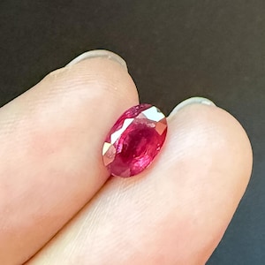 May include: A vibrant, oval-cut ruby gemstone with a deep red hue and multiple facets that reflect light. The stone displays a polished, translucent surface and is held between two fingers against a dark, solid background.