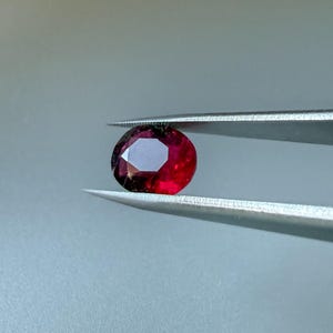May include: A faceted, oval-shaped gemstone with a deep, vibrant red color held between the tips of a pair of silver-toned metal tweezers. The stone displays multiple geometric facets that reflect light, creating a bright, polished appearance.