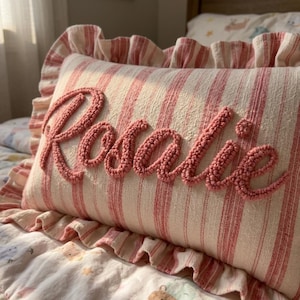 May include: A rectangular throw cushion featuring a cream and red striped pattern with a ruffled border. The name Rosalie is embroidered in a raised, textured, pink yarn script across the centre of the cushion, creating a tactile surface.