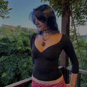Puede incluir: Una blusa negra de manga tres cuartos con un escote en V profundo y un delicado borde de encaje. La prenda se combina con pantalones de rayas horizontales rojas y blancas con cintura de cordón. Se lleva un collar con una gran piedra roja ovalada.