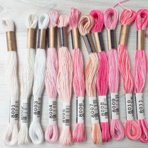 May include: Eleven skeins of embroidery floss arranged in a row, featuring a gradient of colors from white to light pink, coral, and deep magenta. Each skein is secured with a paper band labeled with the brand name Seasons and numbers 8001 to 8011.