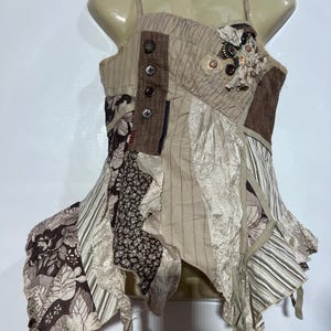Könnte beinhalten: Ein ärmelloses Oberteil mit einem Patchwork-Design aus beigen, braunen und cremefarbenen Stoffen. Das Kleidungsstück weist verschiedene Texturen, Blumen- und Streifenmuster, Zierknöpfe und geraffte Stoffakzente sowie einen asymmetrischen Saum auf.