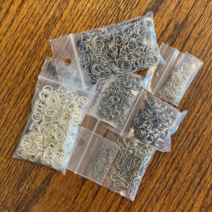 May include: Assortment of silver-colored metal jump rings and clasps in clear plastic bags. The bags are arranged on a wooden surface. The jump rings vary in size and quantity, suitable for jewelry making or crafts.
