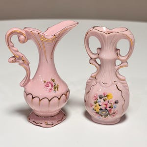 May include: Two small pink ceramic vases with gold trim. The left vase features a single rose design and a curved handle. The right vase has two handles and a floral bouquet pattern. Both pieces have scalloped bases and gold-painted accents.