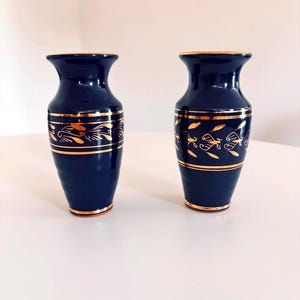 Op de afbeelding: Een paar bijpassende donkerblauwe keramische vazen met een uitlopende rand en een taps toelopende voet. Elke vaas is versierd met horizontale gouden banden en een centraal herhalend goudkleurig bladmotief rond het midden.