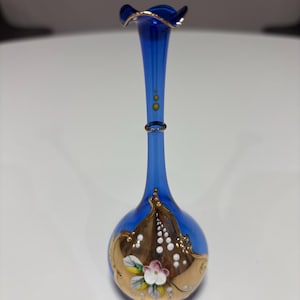 May include: A cobalt blue glass bud vase with a long, slender neck and a ruffled rim. The bulbous base features a gold-painted decorative panel adorned with a hand-painted floral motif in white, pink, and green, accented by small white raised dots.