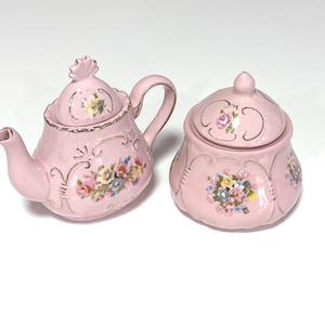 Puede incluir: Una tetera y un azucarero a juego de cerámica rosa claro con tapas. Ambas piezas presentan relieves decorativos, bordes dorados y calcomanías florales coloridas que representan pequeños ramos de rosas y otras flores en el frente.