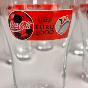 May include: A clear glass featuring a red horizontal label with the Coca-Cola logo, a soccer ball graphic, and the text UEFA EURO 2000. The design includes a stylized colorful emblem next to the text, all printed on the front of the drinking glass.