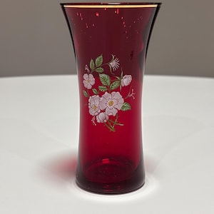 Könnte beinhalten: Eine tief rubinrote Glasvase mit ausgestelltem Rand und konischem, zylindrischem Körper. Auf der Vorderseite befindet sich ein detailliertes Blumenmotiv, das hellrosa Wildrosen mit grünen Blättern und Stängeln auf der glatten Glasoberfläche zeigt.