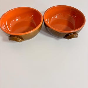 May include: Two round ceramic bowls with a glossy orange interior and a dark brown exterior. Each bowl features a small, curved handle on the side. The bowls have a smooth, reflective finish and are positioned side by side on a plain surface.