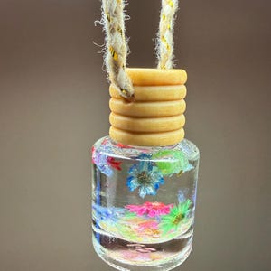 Könnte beinhalten: Eine kleine, durchsichtige Glasflasche, gefüllt mit einer klaren Flüssigkeit und verschiedenen bunten Trockenblumen, darunter blaue, rosa und grüne Blütenblätter. Die Flasche hat einen gerippten, hellen Holzverschluss und hängt an einem dicken Seil.
